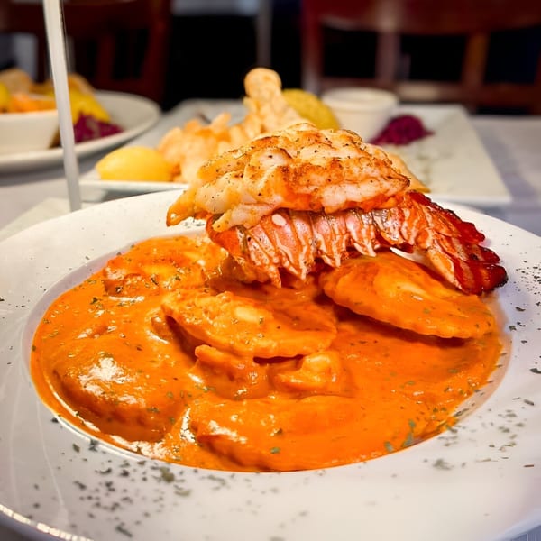 Lobster tail atop ravioli in a vibrant orange sauce, garnished with herbs, at Assado Portuguese Steakhouse.