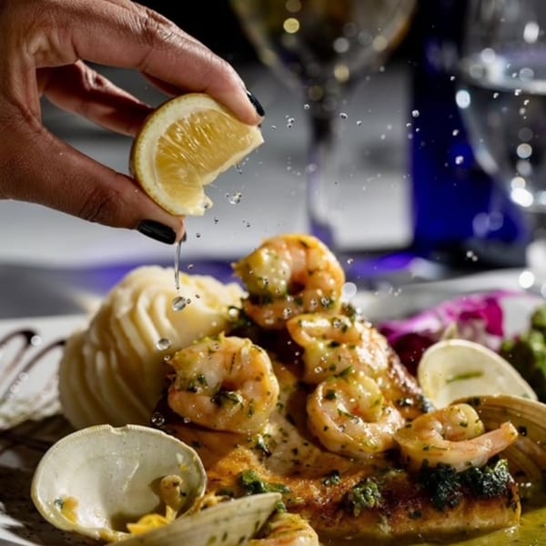A hand squeezes a lemon over a Portuguese seafood dish with shrimp, clams, and mashed potatoes.