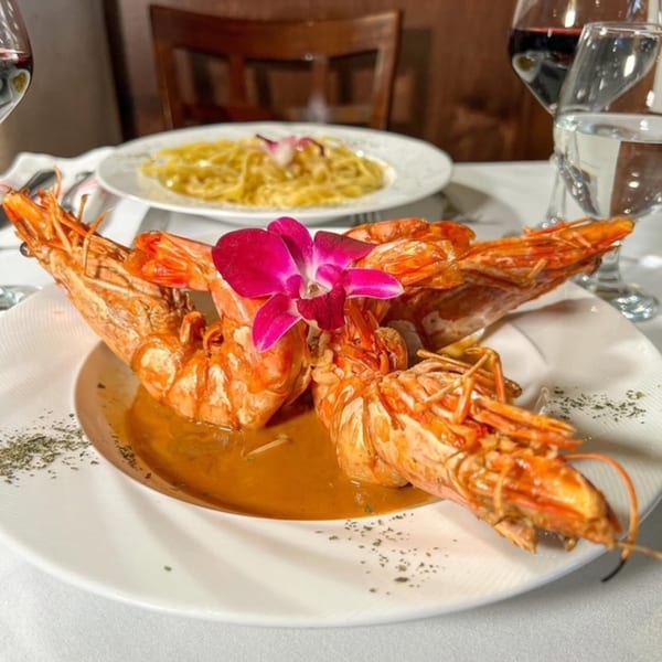 Large grilled prawns in a rich sauce, garnished with an orchid, alongside pasta and wine glasses.